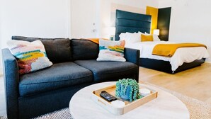 A modern hotel room with a dark grey sofa, a round coffee table with a remote control and a plant, and a bed with a yellow blanket.