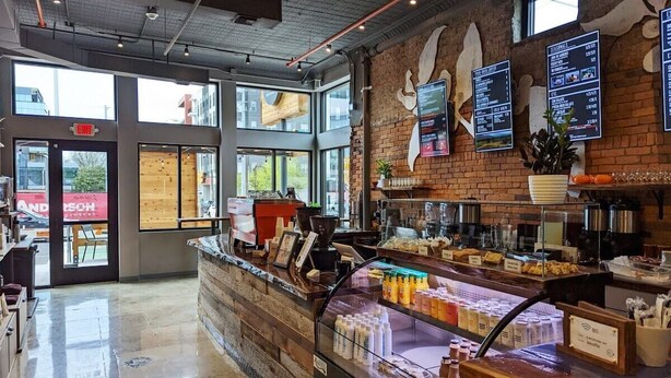 A modern cafe interior with a brick wall, a glass display case with beverages, and a menu board.