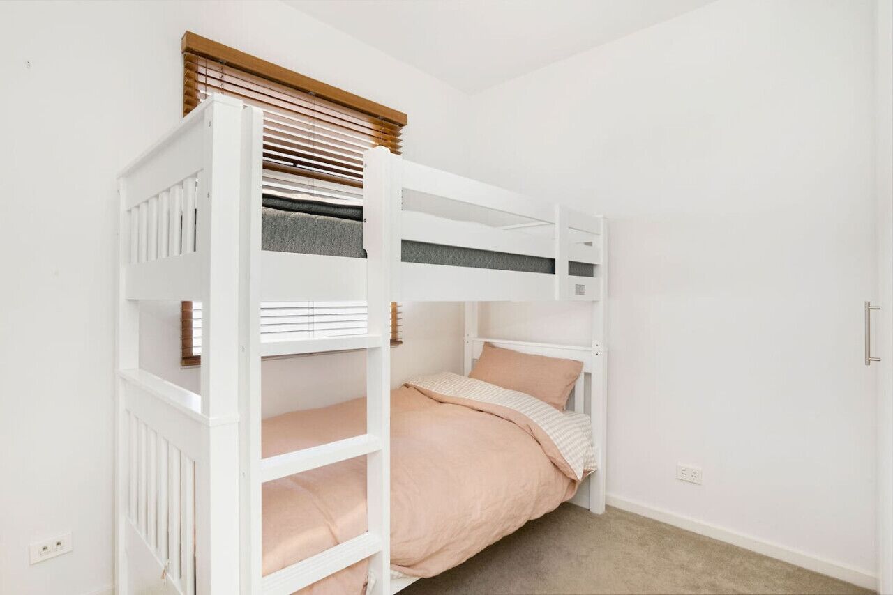 Un lit superposé avec une tête de lit en bois, un lit inférieur recouvert d’une couverture rose et un mur blanc avec une seule prise électrique.
