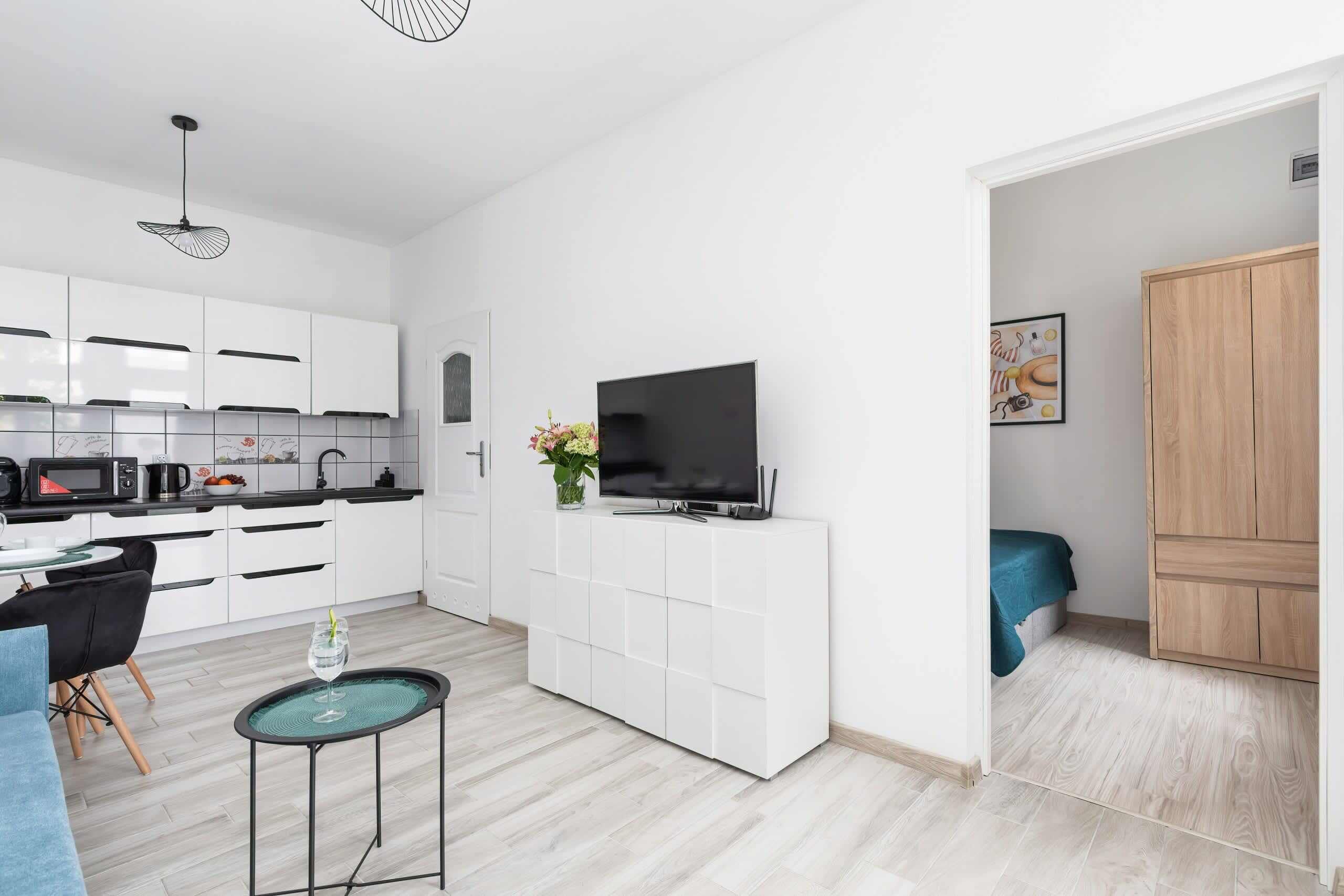 Eine moderne Küche mit weißen Schränken, einem schwarzen Fernseher auf einem weißen Schrank, einem Essbereich mit einer blauen Couch und einem kleinen runden Tisch mit einem Glas.