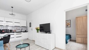 Une cuisine moderne avec des meubles blancs, un téléviseur noir posé sur un meuble blanc, un coin repas avec un canapé bleu et une petite table ronde avec un plateau en verre.