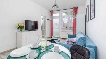 Ein modernes Wohnzimmer mit Fernseher, Esstisch gedeckt mit Tellern und Tassen, einer Couch und einem Fenster mit Vorhängen.