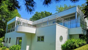 A modern two-story house with a balcony, a white picket fence, and lush greenery. - Maison Moderne en Bois vue mer (Carantec)