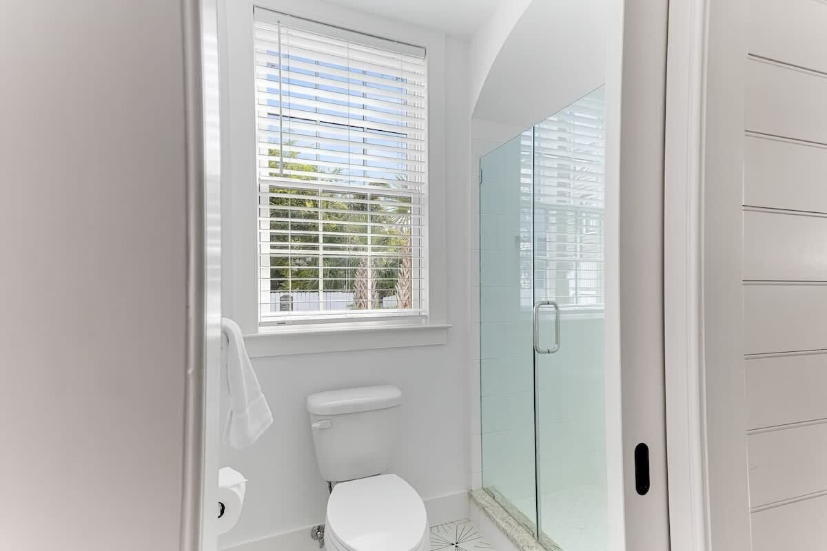 Ein Badezimmer mit Toilette, einem Fenster mit Jalousien und einer Duschkabine aus Glas.