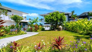 Un jardin bien entretenu, avec un chemin menant à une villa moderne, entourée d’une végétation luxuriante et de plantes tropicales.