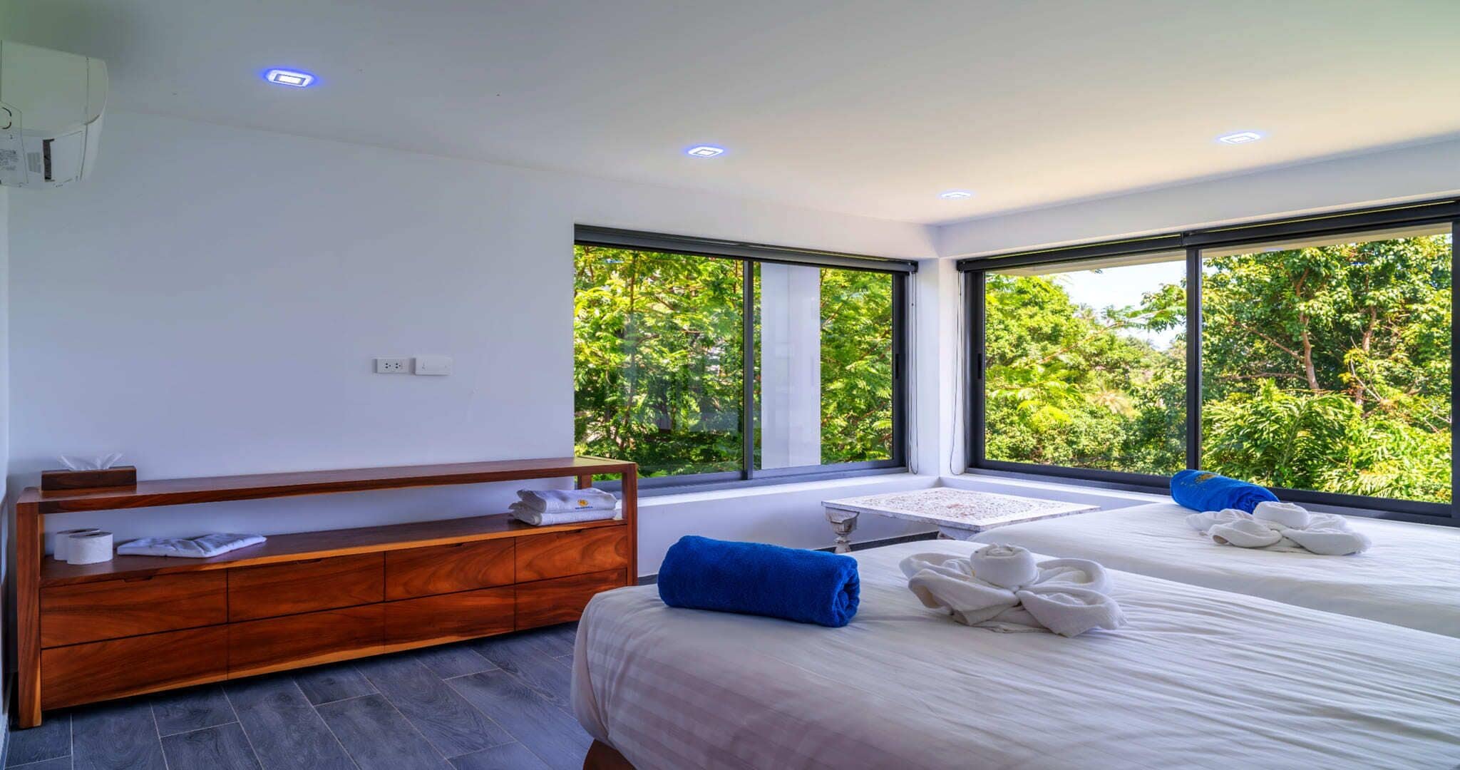 Een moderne hotelkamer met twee bedden, een houten dressoir en grote ramen met uitzicht op weelderige begroeiing.