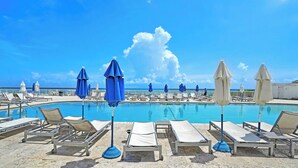 A poolside area with lounge chairs and umbrellas, a clear blue sky, and a calm sea in the background. - Studio with Beach Access and Panoramic City View Miami! (Sunny Isles Beach)