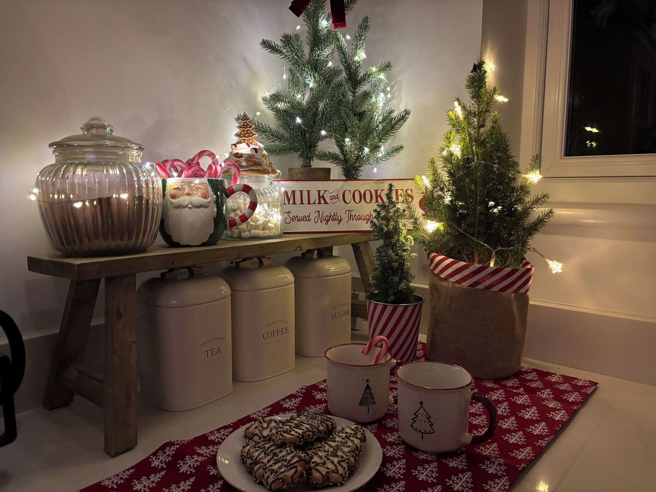 Un rincón de cocina acogedor con una mesa de madera que exhibe varias tazas, un pequeño árbol de Navidad y un letrero que dice 'Se sirven leche y galletas todas las noches'.