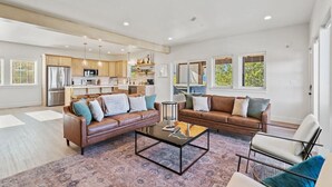 Un amplio espacio de sala con un sofá de cuero marrón, una mesa de centro y una silla. Se ve una cocina al fondo con armarios de madera y un refrigerador.