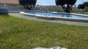 Zona de piscina con cielo despejado, dos arbustos cuidados y un primer plano con césped, una manta y un bolso.