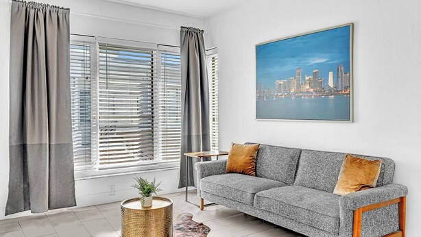 A modern living room with a grey sofa, a cityscape painting, and a view of a city skyline through large windows. - Garden Suites-Hosted by Sweetstay (Miami Beach)