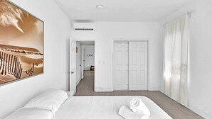 A hotel room with a bed, a painting of a beach scene, a white wardrobe, and a door leading to another room. - Garden Suites-Hosted by Sweetstay (Miami Beach)