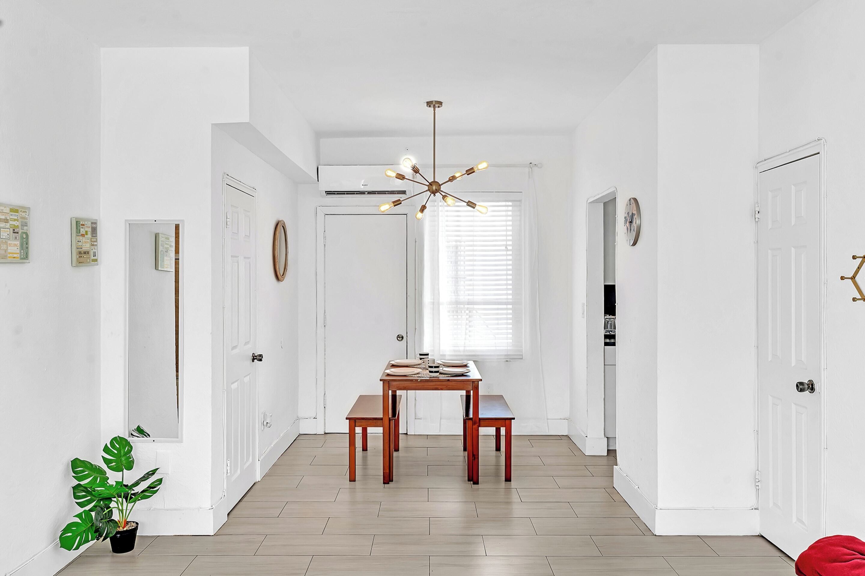 A hallway with a wooden floor, white walls, and a dining table with chairs. There is a potted plant on the left and a red cushion on the right.
