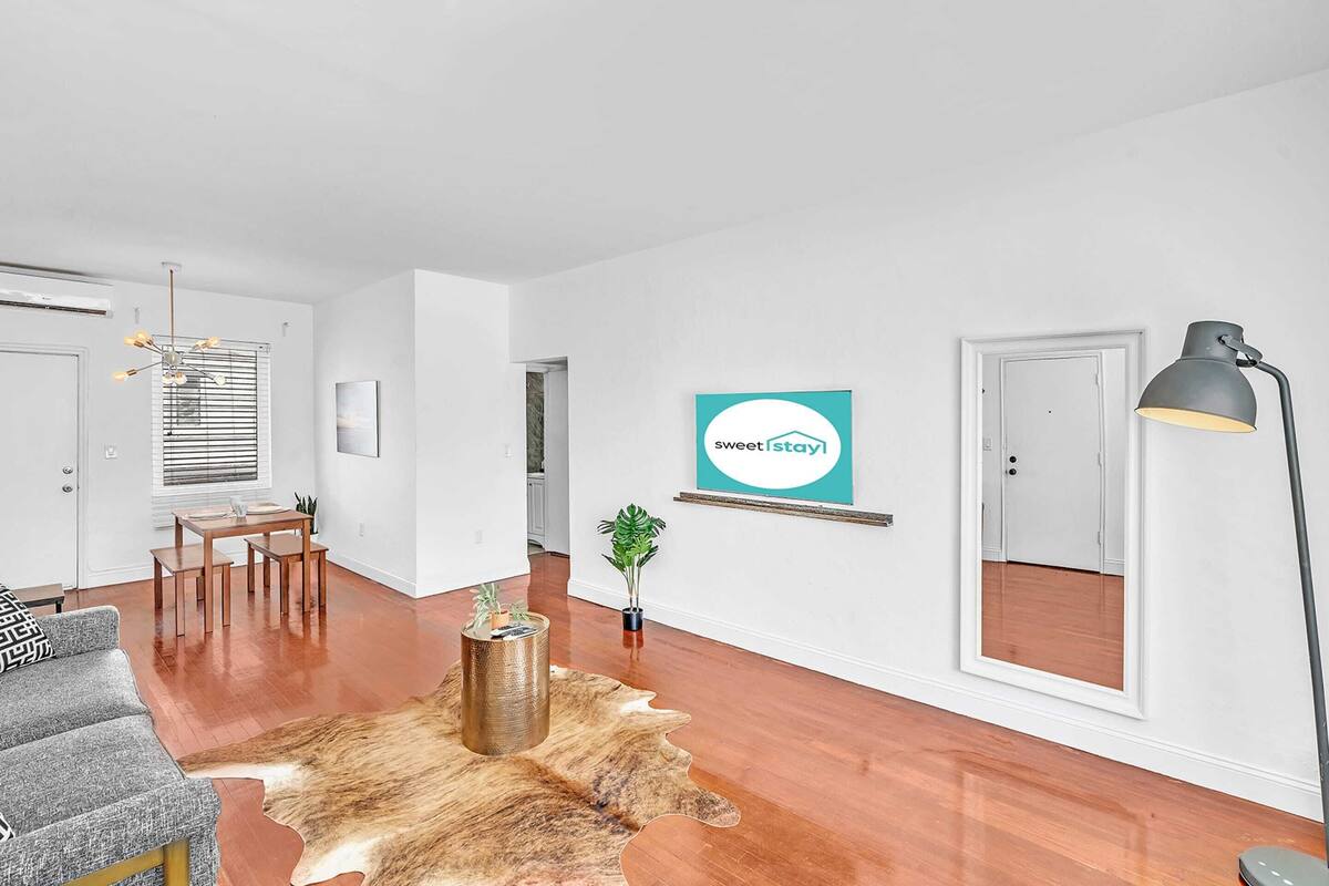 A modern living room with a wooden floor, a grey sofa, a coffee table, a rug, a potted plant, and a wall-mounted sign.