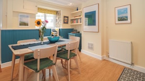 A dining area with a wooden table, chairs, a vase with flowers, and a bookshelf. - Holly Cottage (Yeovil)
