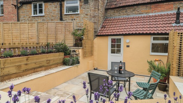 A patio with a table and chairs, surrounded by a garden with purple flowers. - Holly Cottage (Yeovil)