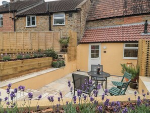 A patio with a table and chairs, surrounded by a garden with purple flowers. - Holly Cottage (Yeovil)