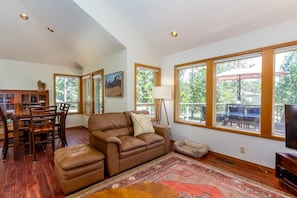 A spacious living room with a brown leather sofa, a dining table with chairs, a TV stand, and large windows offering a view of the outdoors. - Pet-Friendly Sunriver Home + SHARC Passes (Sunriver)