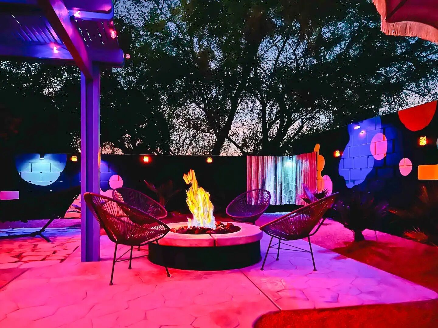Un área de estar al aire libre con un hogar de fuego, rodeada de sillas, bajo una pérgola con luces colgantes.
