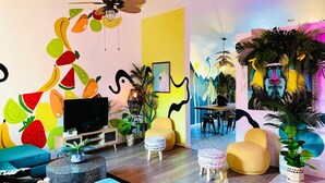 Una sala de estar vibrante con una televisión, un cuadro de pared con frutas coloridas, un ventilador de techo y un comedor con mesa y sillas.