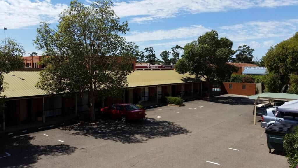 A parking lot with a building in the background, a red car parked, and trees around.