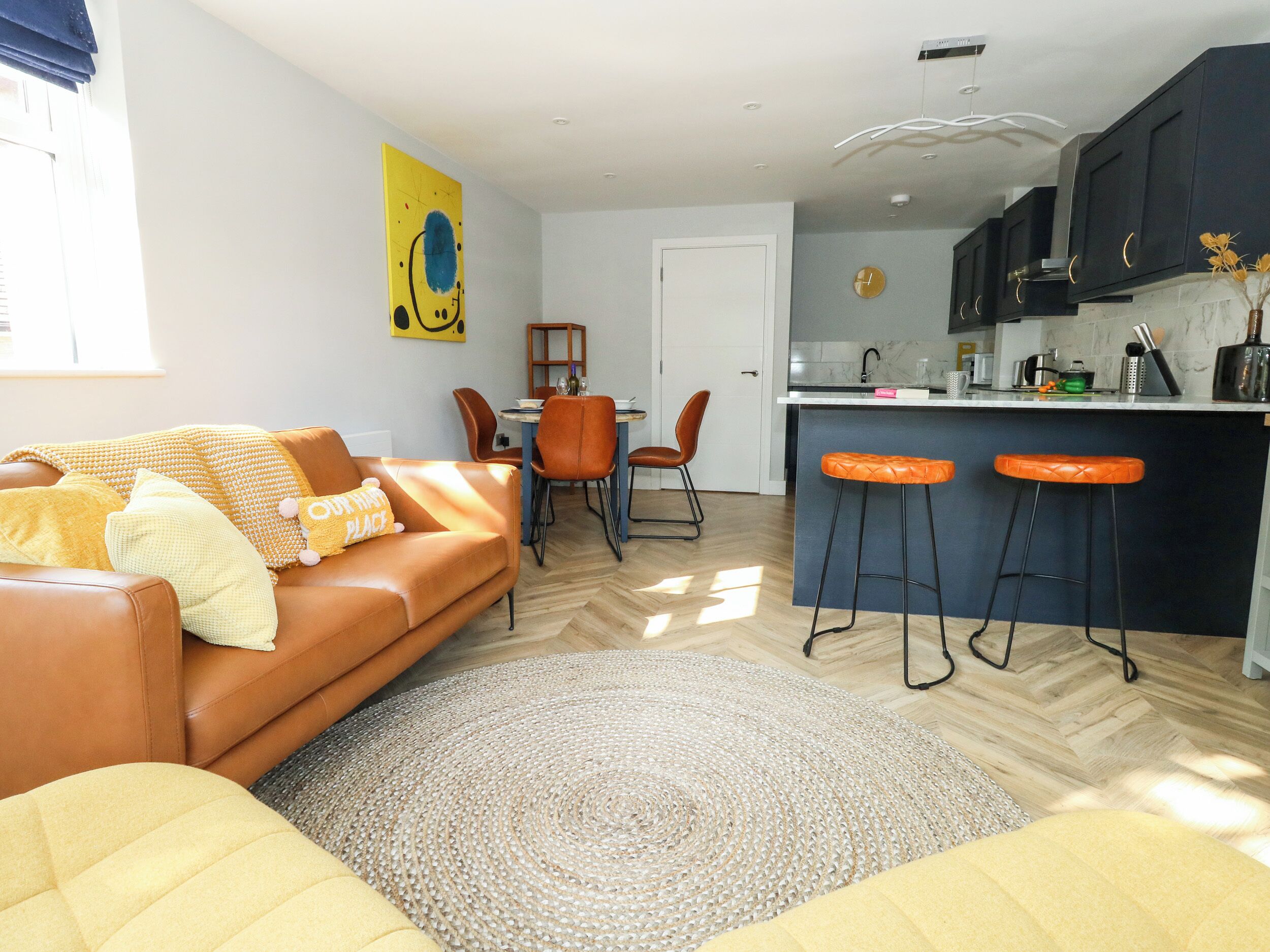 A modern living room with a brown sofa, a dining table with orange chairs, and a kitchen area with dark cabinets and a bar stool.