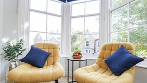 Two yellow armchairs with blue cushions, a small table with a fruit bowl, and a potted plant, all in a room with large windows and a view of buildings outside. - Flat 6, Thornlow Heights (Weymouth)