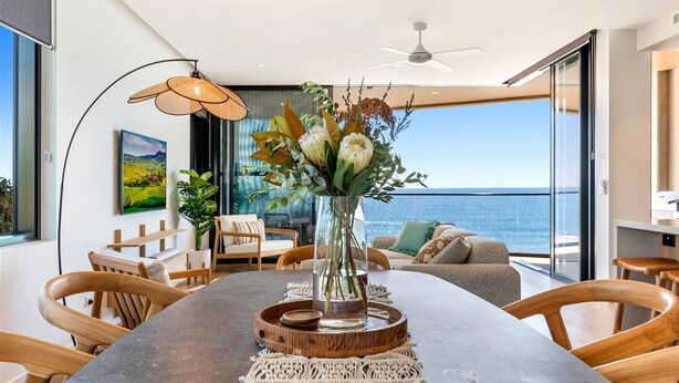 A modern dining area with a large table, wooden chairs, a vase of flowers, and a view of the ocean. - Absolute Beachfront Luxury Apartment (Coolum Beach)