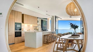 A modern kitchen with wooden cabinets, a central island, and a dining area with a view of the sea. - Absolute Beachfront Luxury Apartment (Coolum Beach)