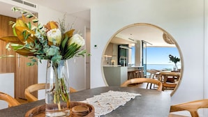 A dining area with a round table, a vase with flowers, and a view of the sea through a large mirror. - Absolute Beachfront Luxury Apartment (Coolum Beach)