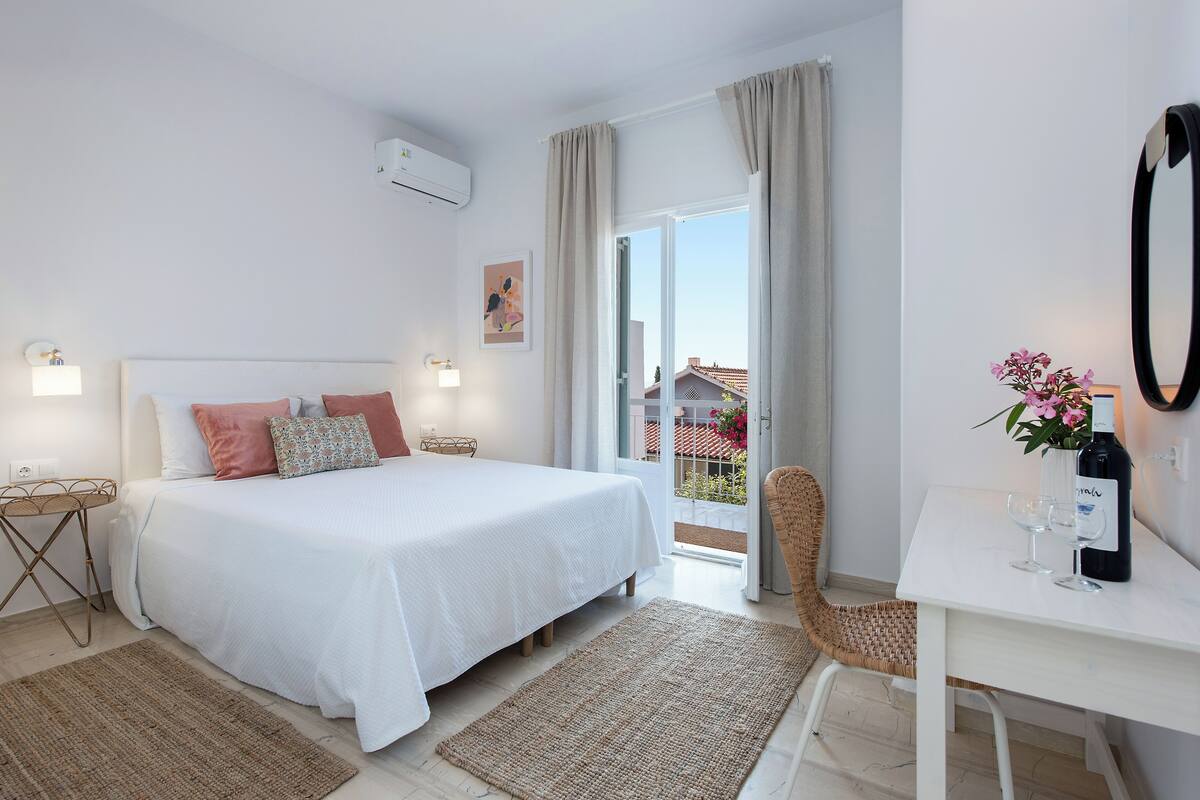A bedroom with a bed, a desk with a bottle and glasses, a chair, and a view of the outside.