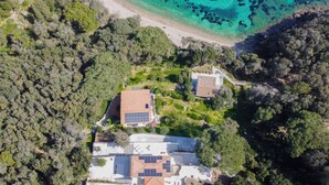 Vue aérienne d’une propriété située sur la côte, avec une plage, une forêt dense et une maison équipée de panneaux solaires.