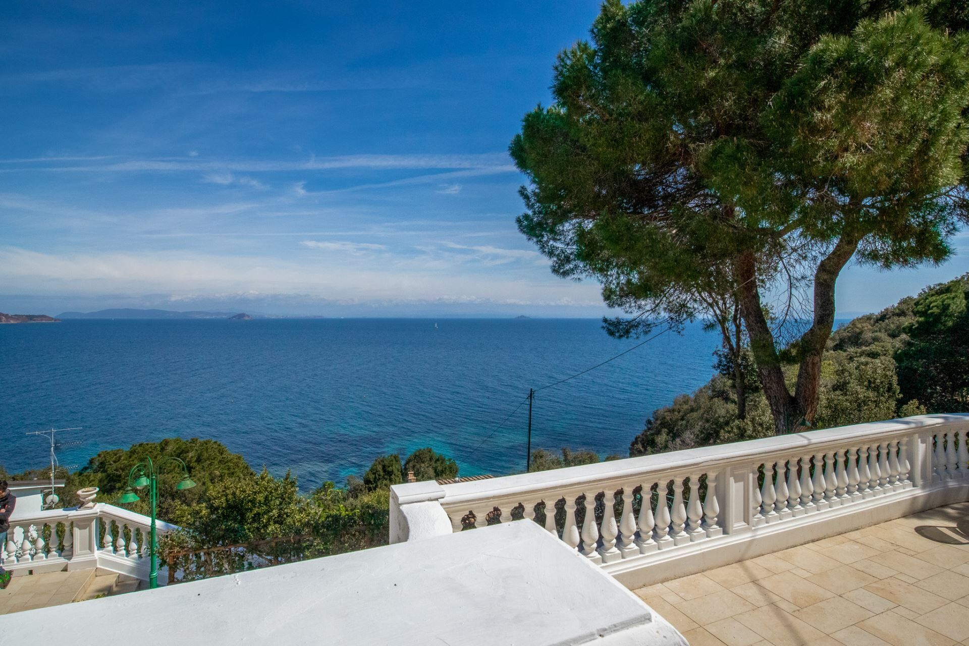 Blick auf die Küste mit blauem Himmel, einem großen Baum und einem weißen Gebäude mit einem Balkon, der auf das Meer blickt.