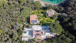 An aerial view of a residential area with two houses, one with solar panels, surrounded by dense greenery and a beach with clear turquoise waters. - Villa Porticciolo has an heartbreaking view. (Rio Marina)