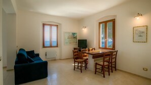 A living room with a blue sofa, a dining table with chairs, a TV, and a window with a view of the sea. - Villa Porticciolo has an heartbreaking view. (Rio Marina)