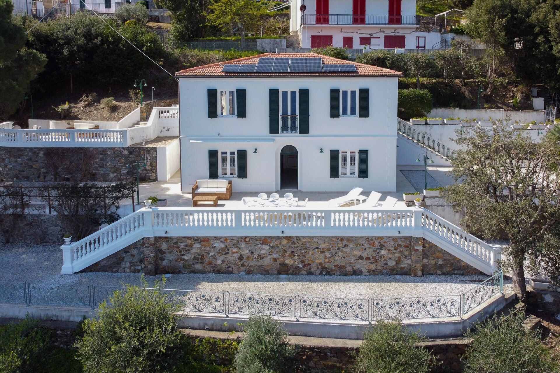 A two-story white building with a stone base, a balcony with white railings, and a solar panel roof.