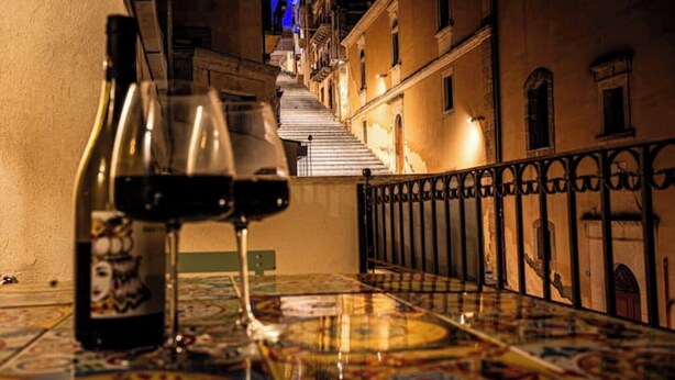 A table with a bottle and glasses of wine, a patterned tablecloth, and a staircase leading to a balcony. - 1 Bedroom Apartment in Caltagirone (Caltagirone)