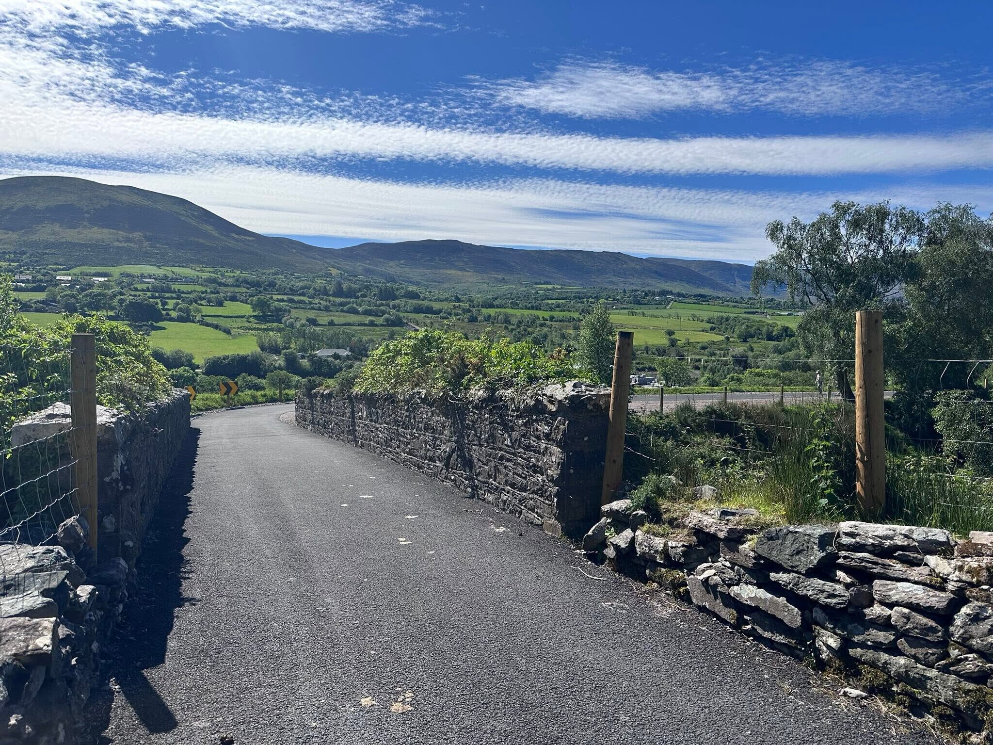A rural road with stone walls and wooden posts, leading through a green valley with hills in the distance.