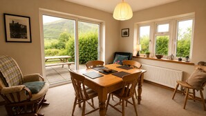 Un espace repas comprenant une table en bois, des chaises, un luminaire suspendu et une vue sur la verdure grâce à de grandes fenêtres.