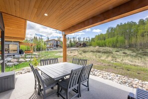 A covered patio with a wooden table and chairs, surrounded by a stone border, overlooking a forested area with houses in the background. - Summit Sky Ranch | Exclusive Amenities | Kid-Friendly Mountain Home (Silverthorne)