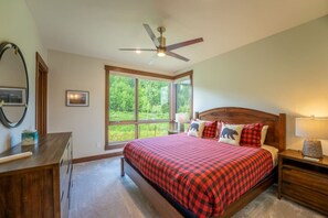 A bedroom with a large window, a bed with a red checkered bedspread, a wooden headboard, a nightstand with a lamp, and a ceiling fan. - Summit Sky Ranch | Exclusive Amenities | Kid-Friendly Mountain Home (Silverthorne)
