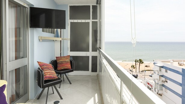 A balcony with a TV, two chairs with colorful cushions, a view of the beach, and a building across the water. - Horizonte - Sea view by HD PROPERTIES (Quarteira)