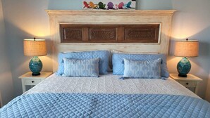 A bed with a quilted headboard, two bedside tables with lamps, and a painting of beach chairs above the headboard.