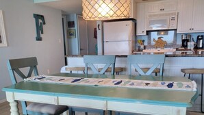 A dining area with a table and chairs, a kitchen in the background, and a pendant light above the table.