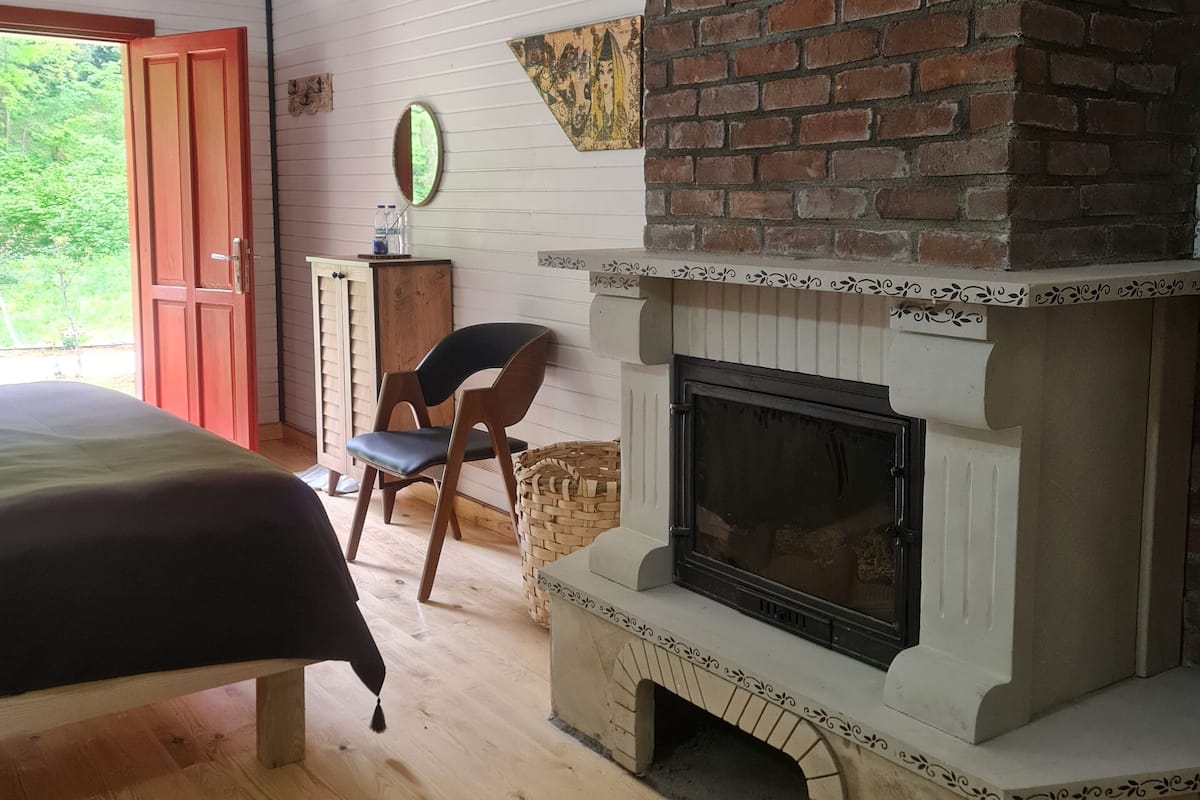 Un dormitorio con chimenea de ladrillo, suelo de madera, una cama, una silla y vistas a la vegetación exterior.