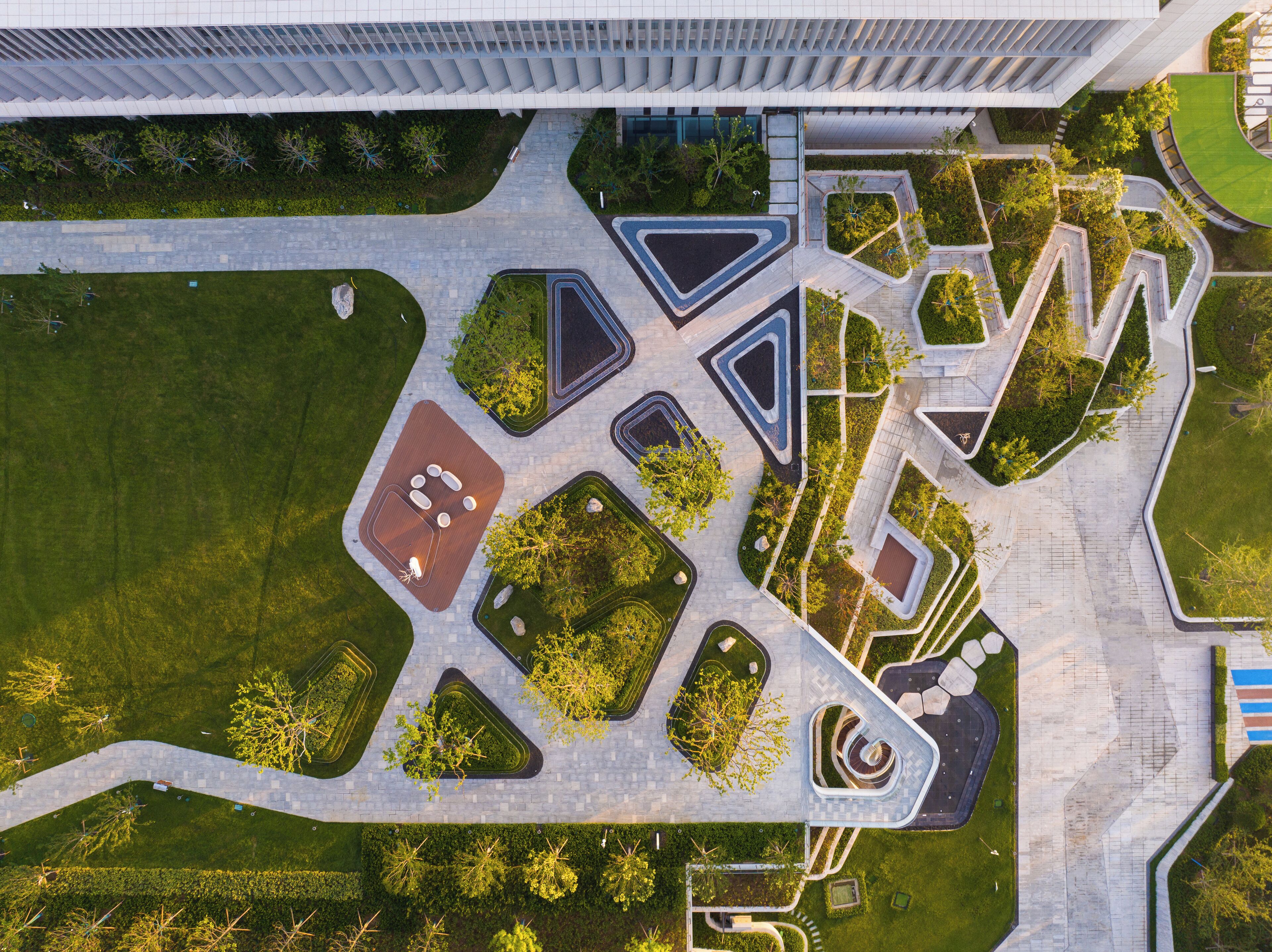 An aerial view of a modern building complex with geometric landscaping, including triangular and rectangular garden areas, and a central spiral feature.