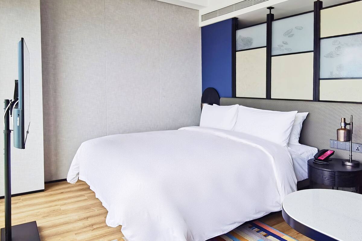 Una habitación de hotel moderna con una cama bien hecha, una mesita de noche con lámpara, una pequeña mesa con una caja azul y una pared con diseño geométrico.