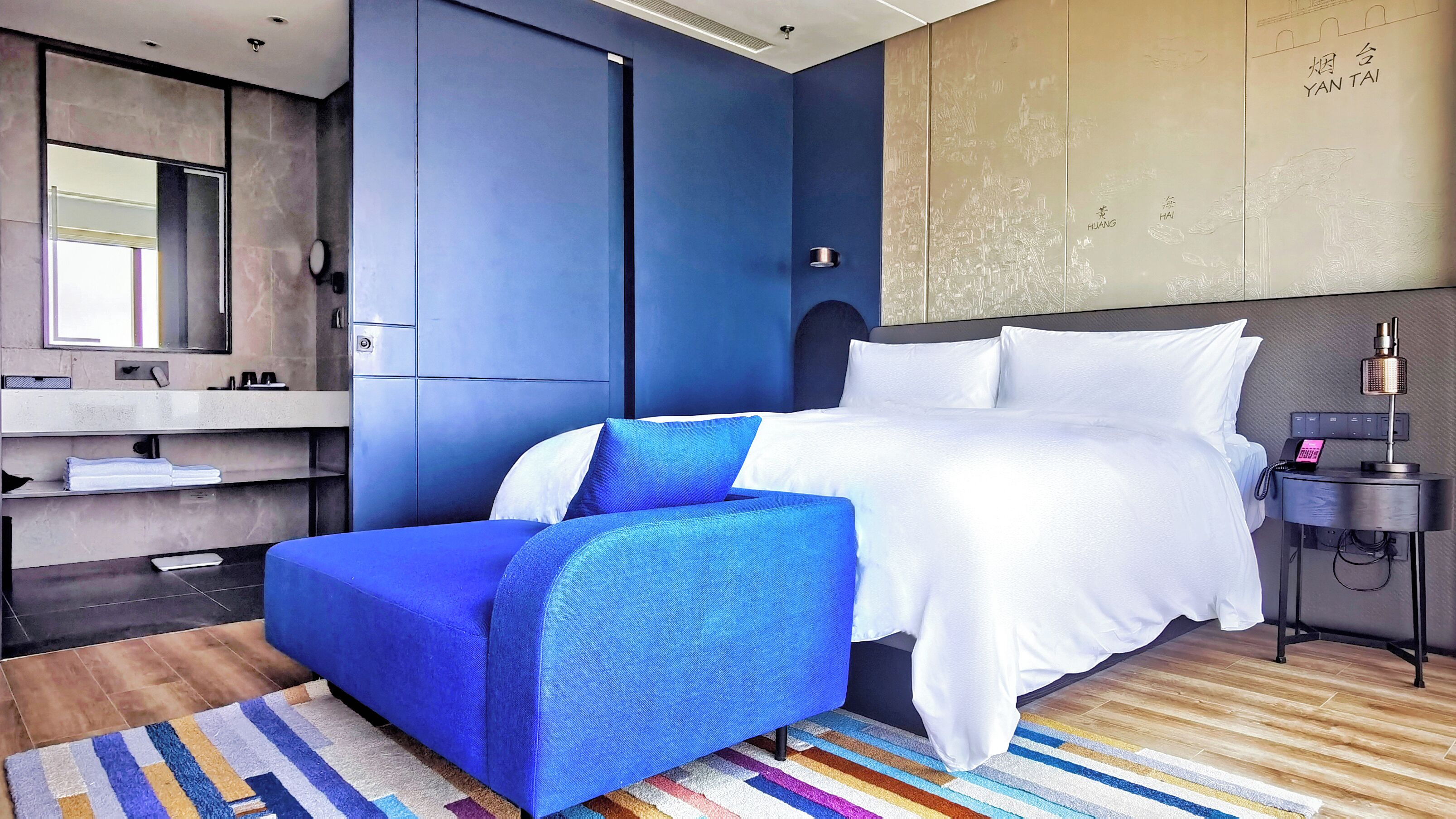 A modern hotel room with a large bed, a blue accent chair, a bedside table with a phone, and a wall-mounted TV.