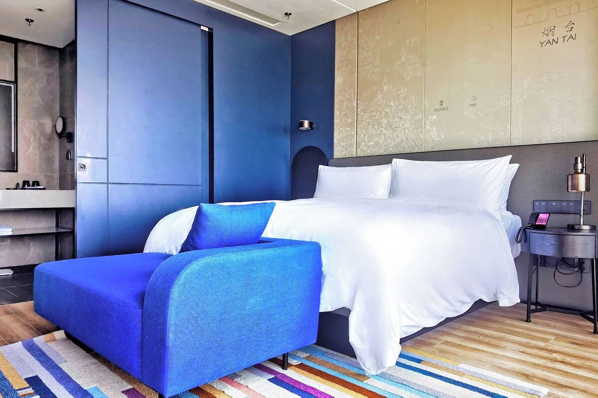 Una habitación de hotel moderna con una cama grande, un sillón azul, una mesita de noche con teléfono y una televisión montada en la pared.