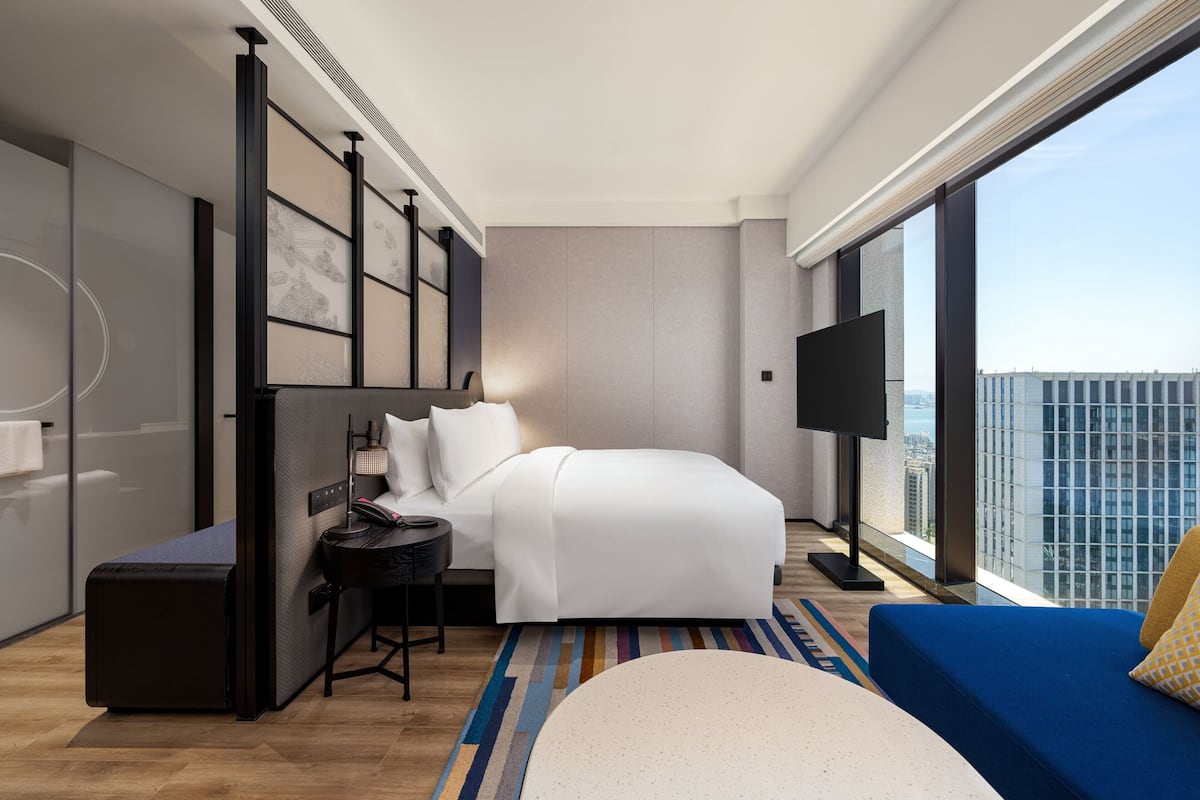 Una habitación de hotel moderna con una cama grande, un escritorio con una silla, un televisor de pantalla plana y un amplio ventanal con vistas a la ciudad.